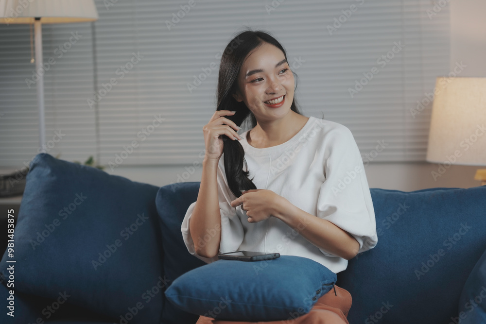Young woman relaxes on her living room sofa, gently touching her hair and smiling, enjoying a peaceful evening at home Young woman relaxes on her living room sofa, gently touching her hair and smiling, enjoying a peaceful evening at home