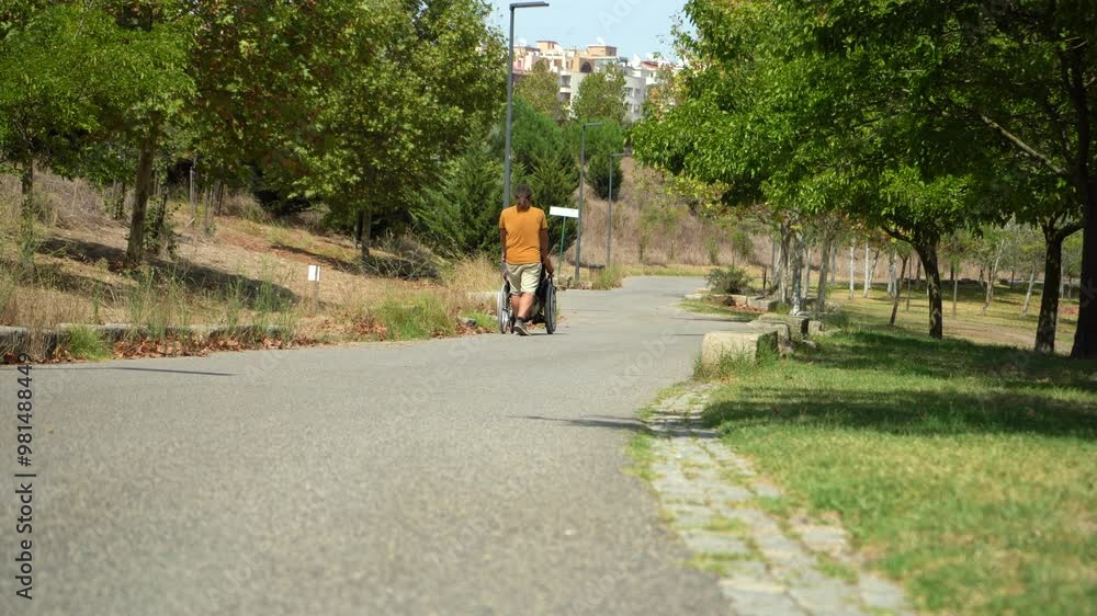 Man Pushing Forward Wheelchair with a Woman on Road in Green Park on Sunny Day. Back View Man Pushing Forward Wheelchair with a Woman on Road in Green Park on Sunny Day. Back View