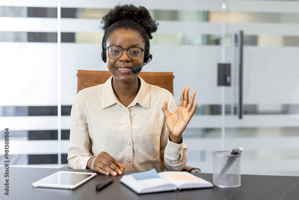 Confident young African American woman using headset for virtual meeting. Smiling and waving, sitting office desk with tablet, notebook, projecting professionalism and friendliness. Confident young African American woman using headset for virtual meeting. Smiling and waving, sitting office desk with tablet, notebook, projecting professionalism and friendliness.