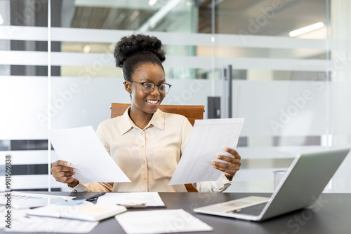 Young african american woman at desk reviewing documents and smiling, working at laptop in modern office setting. Focus on productivity, business analysis, and professional success