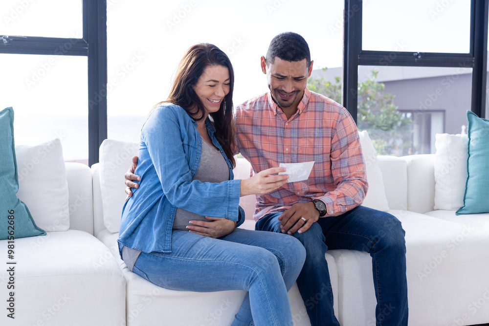 At home, Pregnant multiracial couple sitting on couch, looking at ultrasound picture and smiling At home, Pregnant multiracial couple sitting on couch, looking at ultrasound picture and smiling