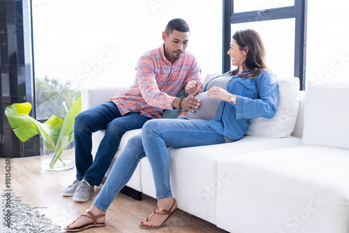 At home, Expecting multiracial couple sitting on couch, man touching woman's pregnant belly lovingly photo