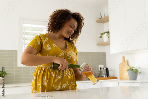 Pouring beer into glass, woman with curly hair enjoying drink in modern kitchen at home