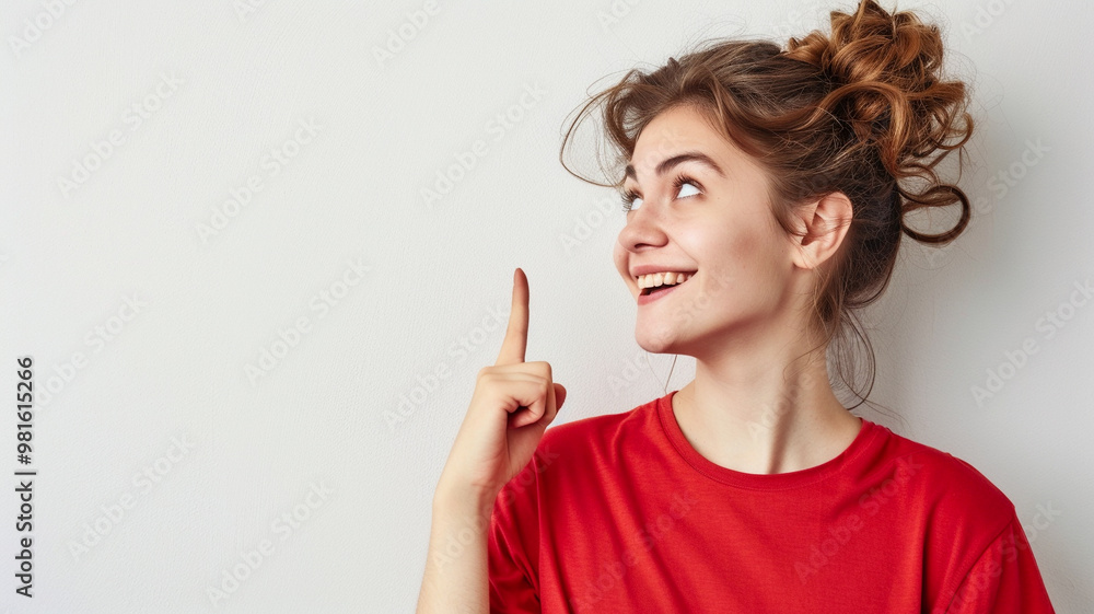 A young woman with her hair in a messy bun smiles brightly, pointing upwards, wearing a red shirt. The image has a light minimalist background A young woman with her hair in a messy bun smiles brightly, pointing upwards, wearing a red shirt. The image has a light minimalist background