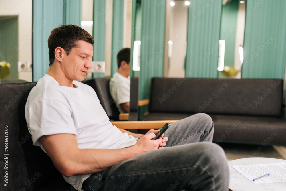 Happy muscular man traveler in white clothes relaxing in hotel lobby using smartphone watching online video or browsing internet. Male tourist resting with phone in lounge area at luxury resort. Happy muscular man traveler in white clothes relaxing in hotel lobby using smartphone watching online video or browsing internet. Male tourist resting with phone in lounge area at luxury resort.