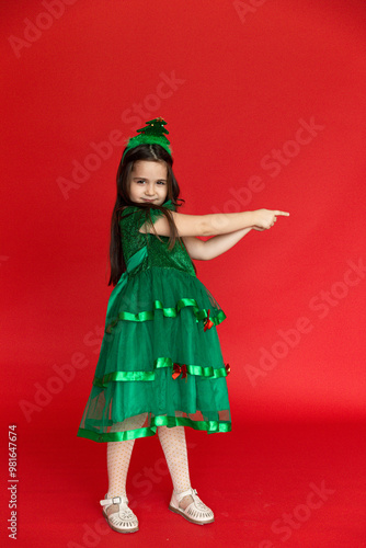 A girl in a festive carnival costume of little Christmas tree pointing at an empty space on a red background. Winter holidays concept. Sale and advertising