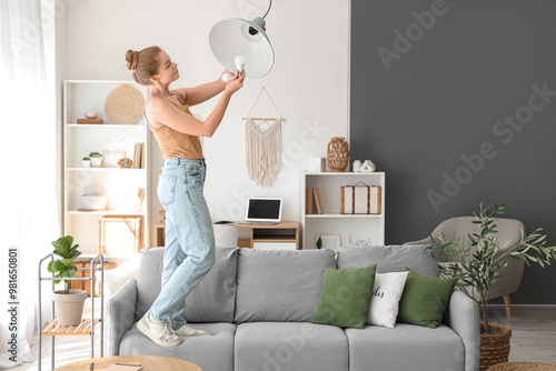 Slika na platnu Young woman on sofa screwing light bulb into hanging lamp at home