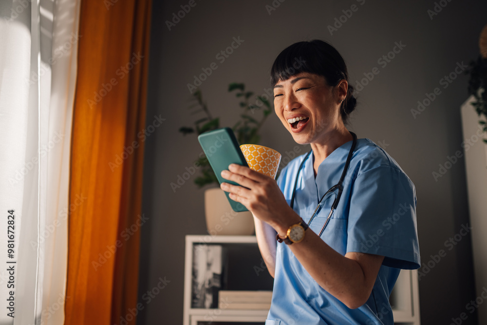 Asian woman nurse standing by window, with phone and yellow patterned mug Asian woman nurse standing by window, with phone and yellow patterned mug
