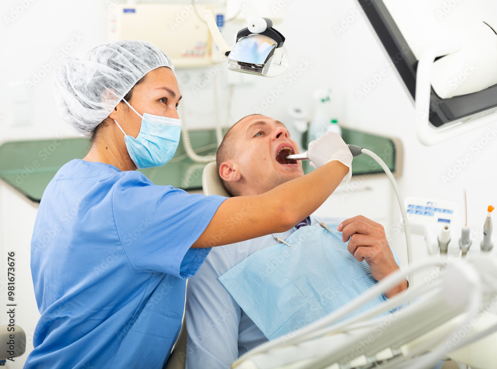 Fototapeta premium Qualified woman dentist in a protective mask working in the clinic conducts an examination of a man patient sitting in a ..dental chair using an apexlocator