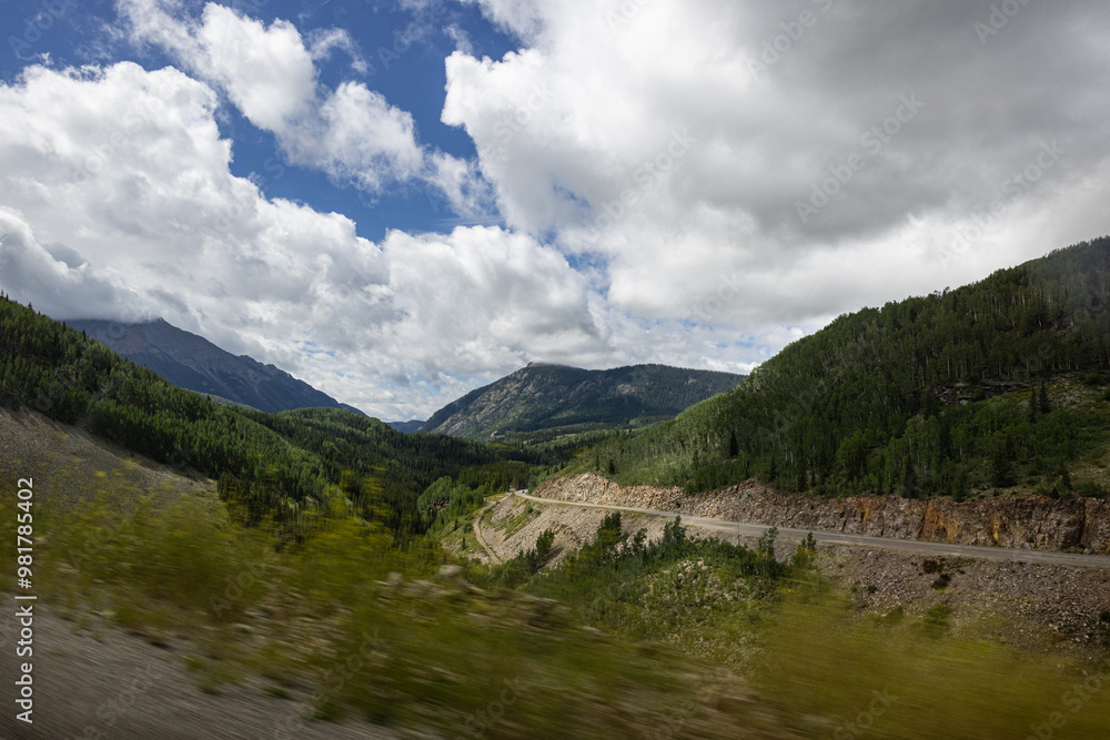 Fototapeta premium Photograph taken from moving vehicle of whinnying road mountainous scenery and vivd cloudy bright blue sky in Colorado