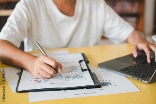 Close-up of a person analyzing financial charts and graphs with a pen and laptop on a yellow desk.