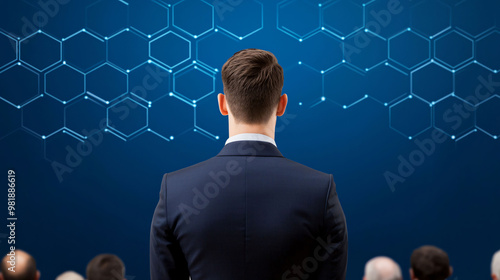 A professional speaker stands confidently in front of large digital display, engaging an audience with captivating presentation. backdrop features modern, abstract design, enhancing atmosphere of inno photo