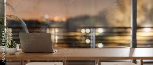 A laptop on a wooden tabletop in a room by the window, offering a dusk view through a balcony.
