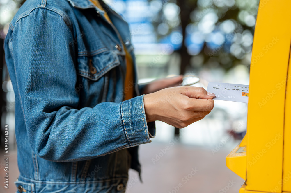 A close-up of a woman in a denim jacket inserting her debit card into an ATM to withdraw some money. A close-up of a woman in a denim jacket inserting her debit card into an ATM to withdraw some money.