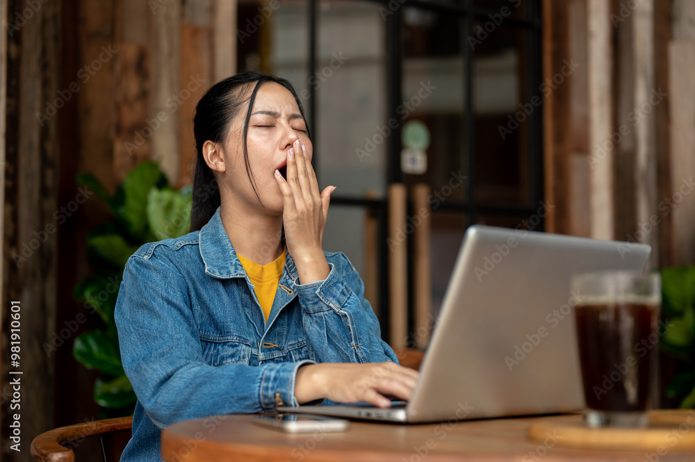 A tired, sleepy Asian woman is yawning, feeling drowsy while working remotely in a coffee shop. A tired, sleepy Asian woman is yawning, feeling drowsy while working remotely in a coffee shop.