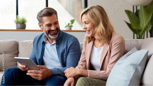 Happy middle-aged Caucasian man and woman using a tablet while sitting on a sofa at home.