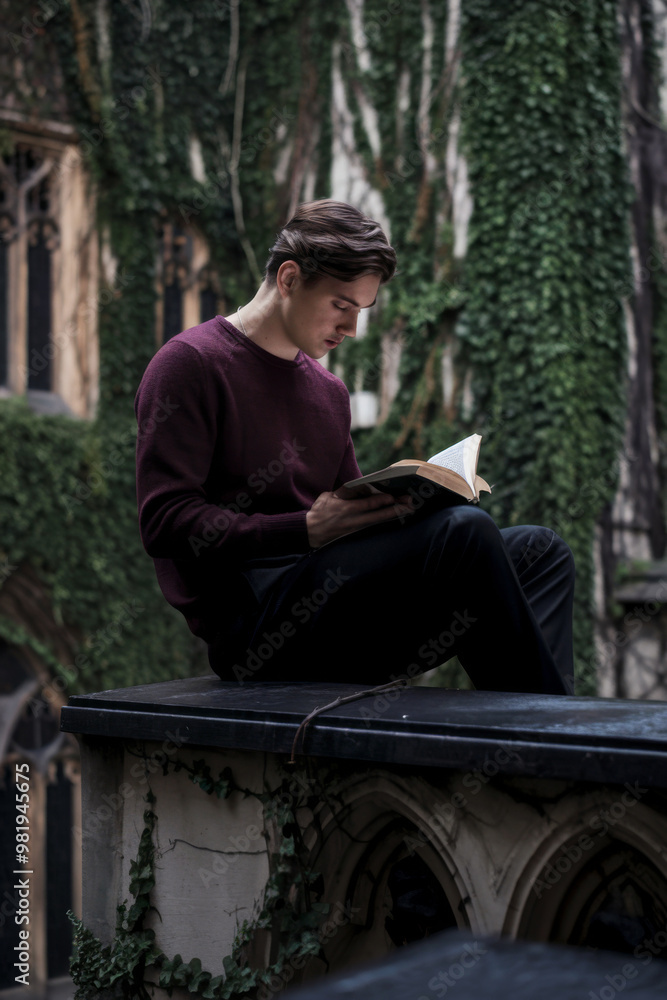 Obraz premium Young man sitting on a ledge reading a book in a old university with ivy-covered walls. Concept of education and quiet contemplation.