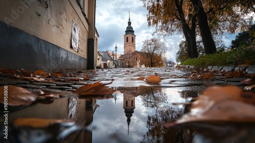 Reflejo de un campanario en un charco de agua en una calle empedrada cubierta de hojas durante el Día de Todos los Santos. El ambiente otoñal y la arquitectura destacan la solemnidad de la imagen. photo