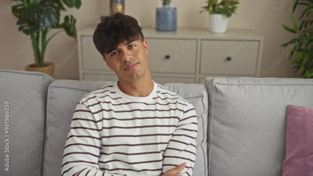 Young handsome man in a striped shirt sitting on a couch in a modern living room at home, smiling and crossing his arms while looking at the camera. Young handsome man in a striped shirt sitting on a couch in a modern living room at home, smiling and crossing his arms while looking at the camera.