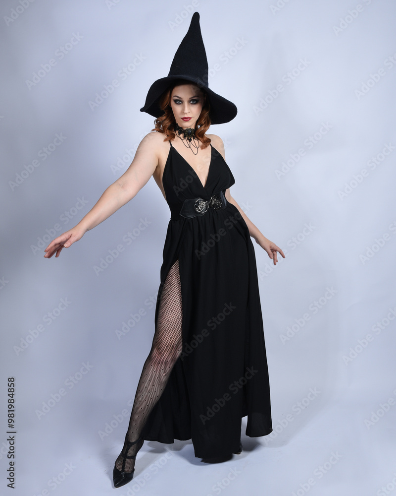 Full length portrait of beautiful female red head model wearing glamorous black witch dress with pointy hat halloween costume. walking pose with gestural arms, isolated figure on studio background Full length portrait of beautiful female red head model wearing glamorous black witch dress with pointy hat halloween costume. walking pose with gestural arms, isolated figure on studio background