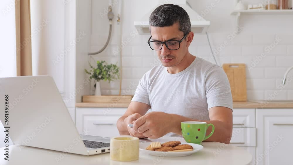 Handsome man freelancer working on online projects using phone and laptop while having serious phone call and focusing on remote work in home kitchen setting Handsome man freelancer working on online projects using phone and laptop while having serious phone call and focusing on remote work in home kitchen setting