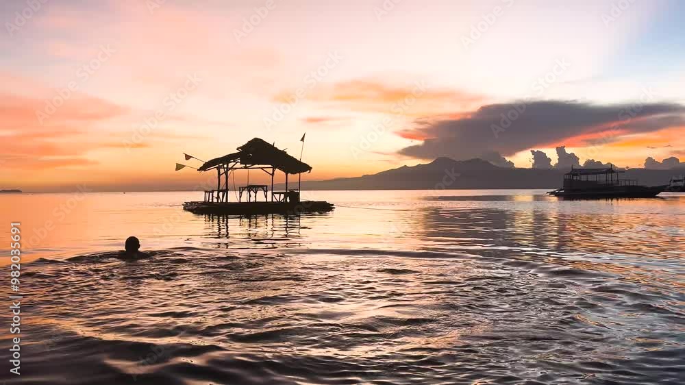 A man dives into a calm ocean and then swims away during a beautiful and colourful sunset on Paliton Beach in Suquijor Island, Philippines. The sky is full of shades of orange. A man dives into a calm ocean and then swims away during a beautiful and colourful sunset on Paliton Beach in Suquijor Island, Philippines. The sky is full of shades of orange.