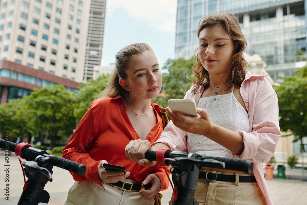 Obraz premium Two young women examining smartphone while standing with electric scooters amidst urban buildings. They seem engaged and curious about their surroundings
