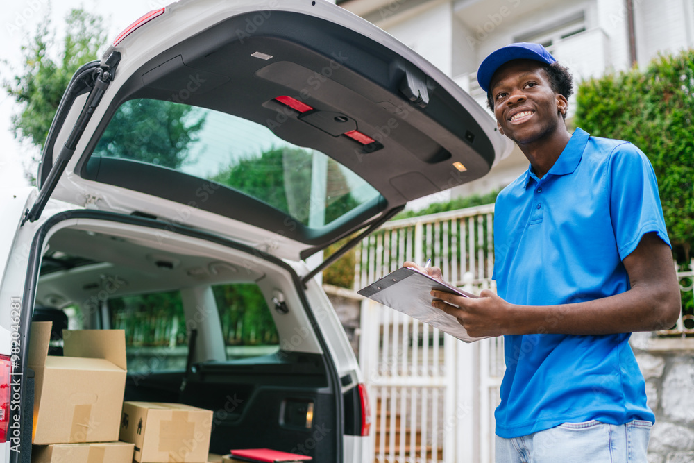 Smiling young delivery man holding clipboard next to car filled with packages. Smiling young delivery man holding clipboard next to car filled with packages.