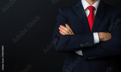 Confident businessman with crossed arms in formal suit and red tie against a dark background, exuding professionalism and authority.