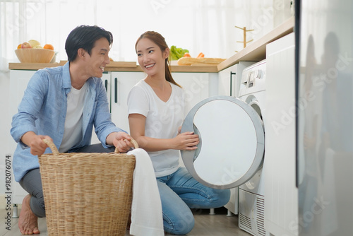 Happy young asian couple doing laundry fold clothes together using a front loading washing machine to wash laundry. Husband and wife during household routine in morning at home.