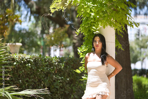 Young, pretty, dark-haired, Latina woman in a white dress, looking at the camera leaning on a marble column with green plant leaves. Concept beauty, fashion, trend, plants, spring. photo