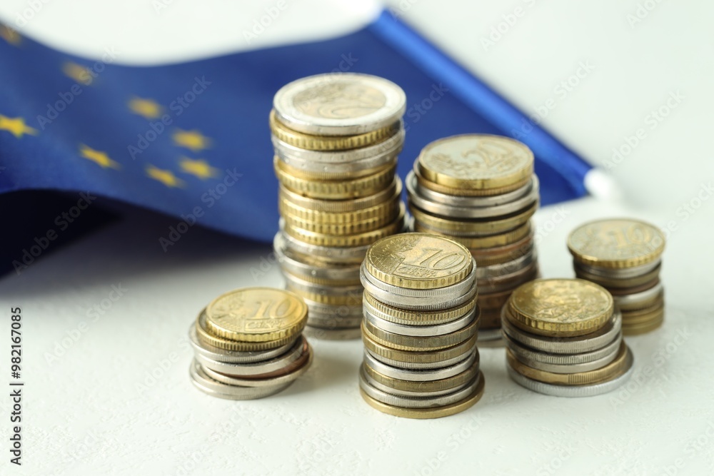 Stacks of euro coins and European Union flag on white textured table, closeup