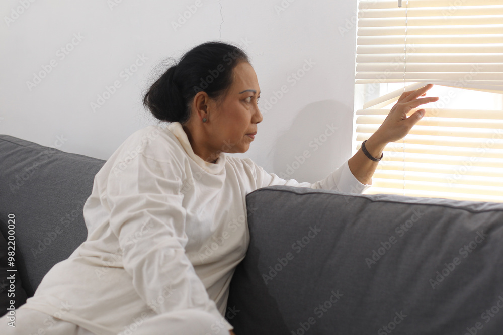 Asian Mature Woman Sitting On Sofa And Peeking Looking Through Window Blinds Asian Mature Woman Sitting On Sofa And Peeking Looking Through Window Blinds