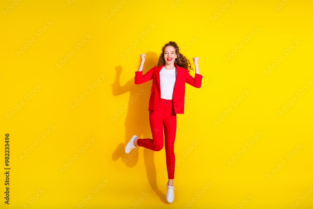 Full size photo of pretty young woman jump raise fists wear red suit isolated on yellow color background Full size photo of pretty young woman jump raise fists wear red suit isolated on yellow color background