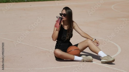 Woman basketball player take a rest during the training. Sitting on basketball court and drinking water.