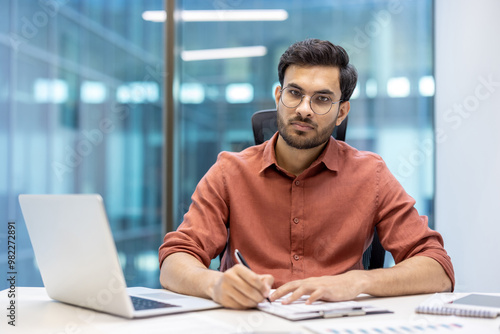 Businessman sitting at office desk, using laptop and writing notes. Professional atmosphere and serious expression illustrate focused work in modern workplace environment.