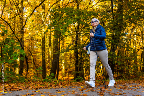 Beautiful middle-aged blonde woman in casual wear practicing Noridc walking on sidewalk in autumn scenery. Side view. Autumnal Nordic walking in city.