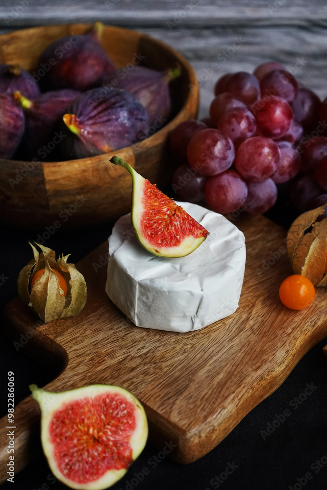 Ripe figs next to Brie cheese on a wooden board, pink grapes and Physalis fruits on a dark background Ripe figs next to Brie cheese on a wooden board, pink grapes and Physalis fruits on a dark background