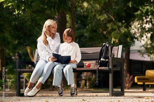 Information in notepad, reading it. Little schoolgirl with her mother are sitting on the bench outdoors