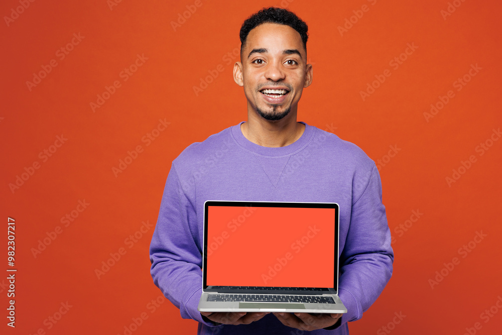 Young IT man of African American ethnicity he wear purple sweatshirt casual clothes hold use work on blank screen laptop pc computer isolated on plain red orange background studio. Lifestyle concept. Young IT man of African American ethnicity he wear purple sweatshirt casual clothes hold use work on blank screen laptop pc computer isolated on plain red orange background studio. Lifestyle concept.