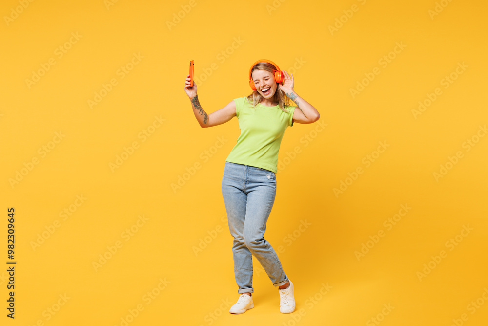Full body young woman she wear green t-shirt casual clothes listen to music in headphones hold in hand use mobile cell phone dance sing isolated on plain yellow orange background. Lifestyle concept. Full body young woman she wear green t-shirt casual clothes listen to music in headphones hold in hand use mobile cell phone dance sing isolated on plain yellow orange background. Lifestyle concept.