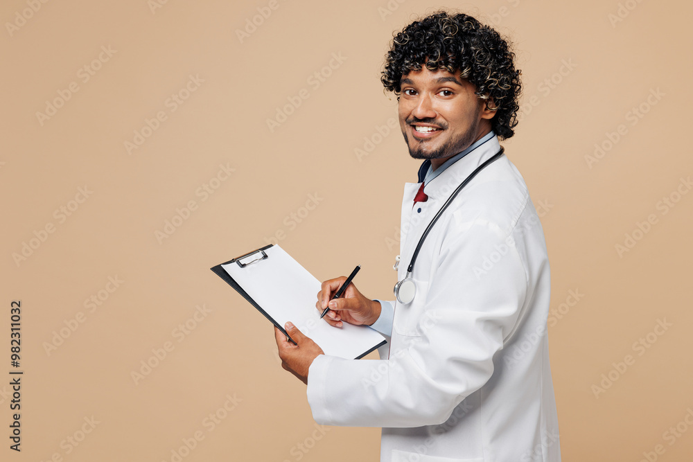 Sideways young happy Indian doctor man in white medical gown suit work in hospital clinic office hold clipboard with patient card isolated on plain beige background studio Healthcare medicine concept Sideways young happy Indian doctor man in white medical gown suit work in hospital clinic office hold clipboard with patient card isolated on plain beige background studio Healthcare medicine concept