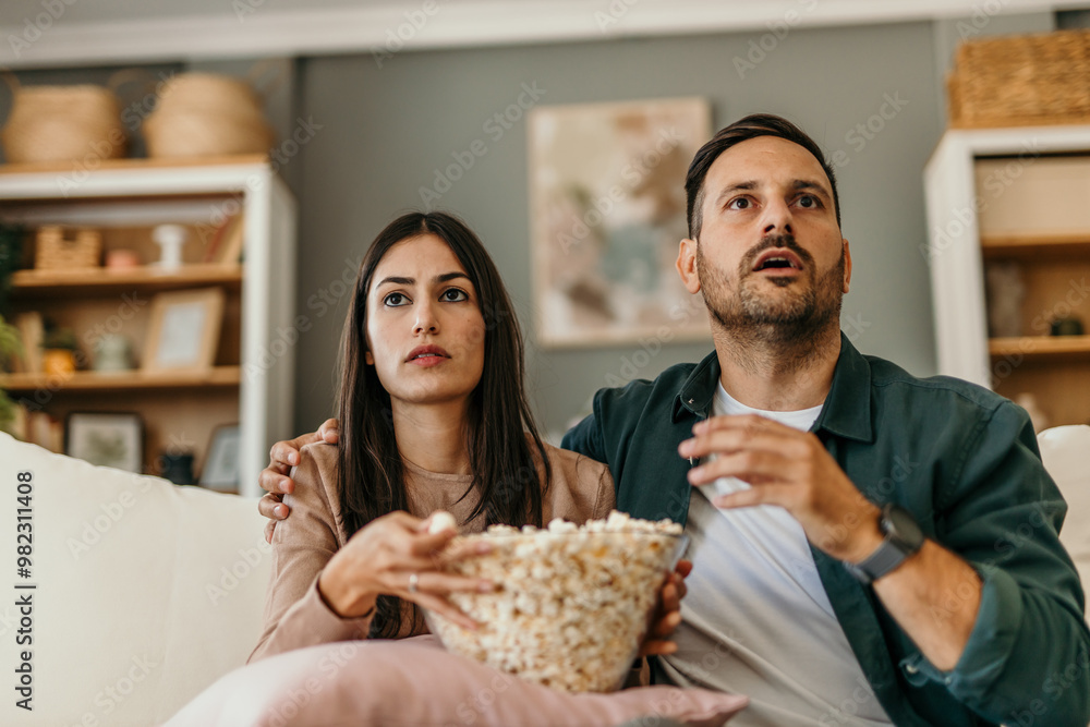 Multiracial husband and wife eating popcorn from a bowl and watching drama movie while chilling on the couch in weekend Multiracial husband and wife eating popcorn from a bowl and watching drama movie while chilling on the couch in weekend