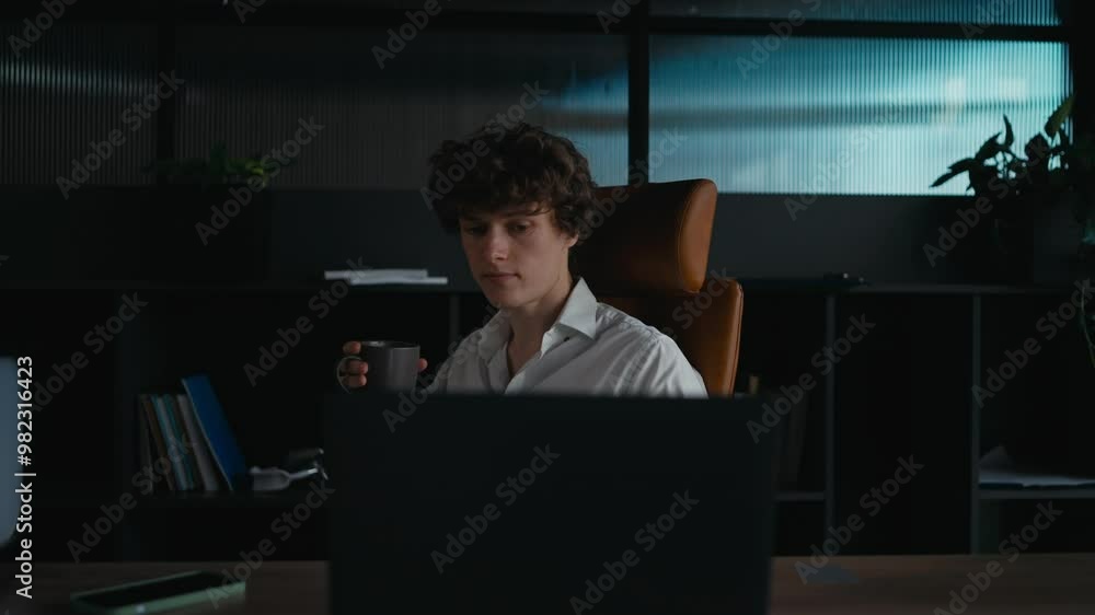 A young guy with curly hair in a white shirt sits at a table and works on a laptop while drinking coffee in the office