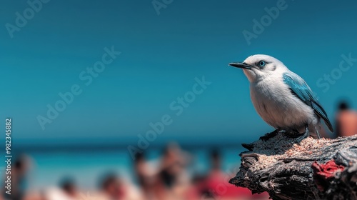 A small bird with blue and white feathers perches calmly on a piece of weathered wood, against a backdrop of a busy beach with blurred figures enjoying the sun. photo