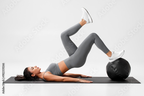 Athletic girl doing glute bridge exercise with med ball on gray background. Fitness woman working out