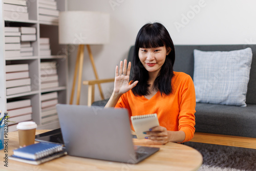 asian woman is sitting on a white couch in a modern home. Her is smiling and looking at her laptop freelance business online concept. 