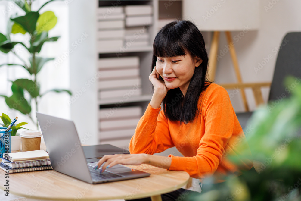 asian woman is sitting on a white couch in a modern home. Her is smiling and looking at her laptop freelance business online concept.
