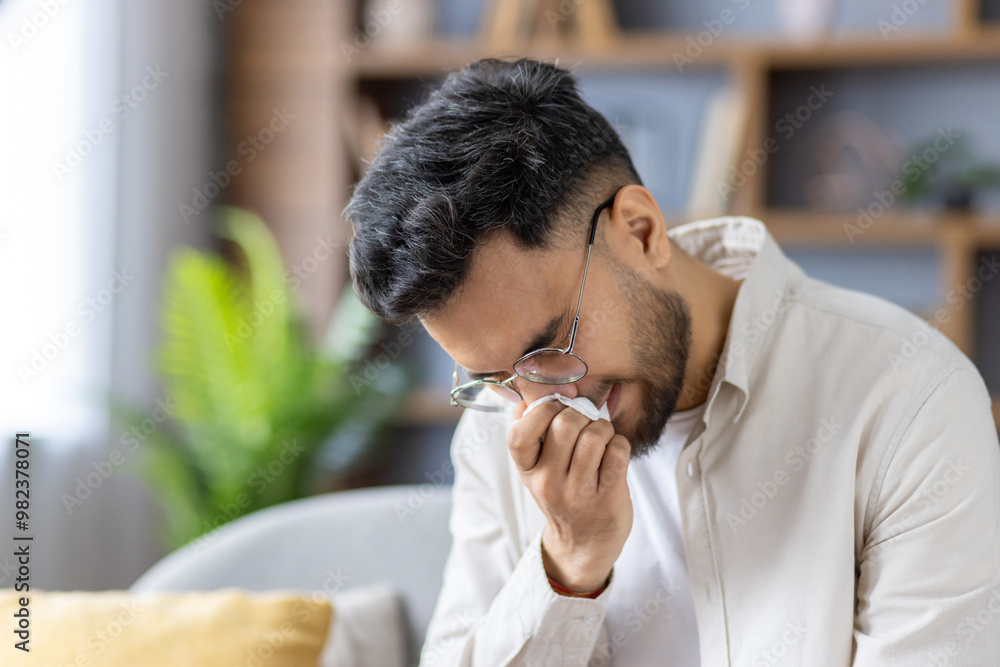 Man experiencing cold symptoms holding tissue to nose while sitting indoors in casual outfit Man experiencing cold symptoms holding tissue to nose while sitting indoors in casual outfit