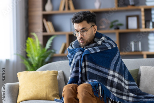 Картината върху платно Man wrapped in blanket sitting on sofa at home looking cold and shivering, tryin